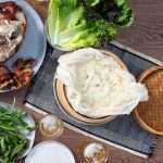 Overhead shot of a steamer basket full of sticky rice alongside a spread of roast pork shoulder, Thai herbs, and lettuces.